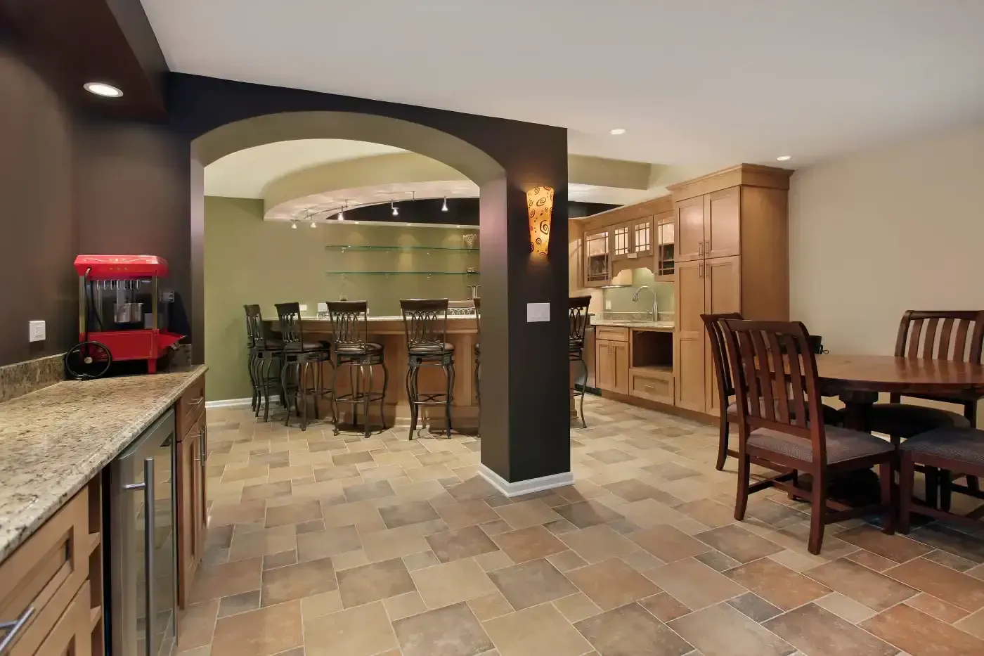 Elegant basement with a beige tiled floor, featuring a wooden bar area with high chairs under a curved archway. A dining table and chairs sit nearby, with warm lighting creating a cozy atmosphere.