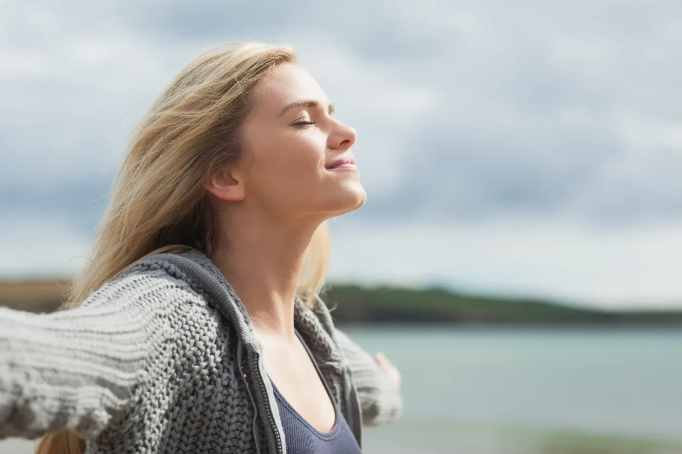 A woman with closed eyes smiles softly, arms outstretched, wearing a gray sweater near a calm sea under a cloudy sky, conveying peace and freedom.