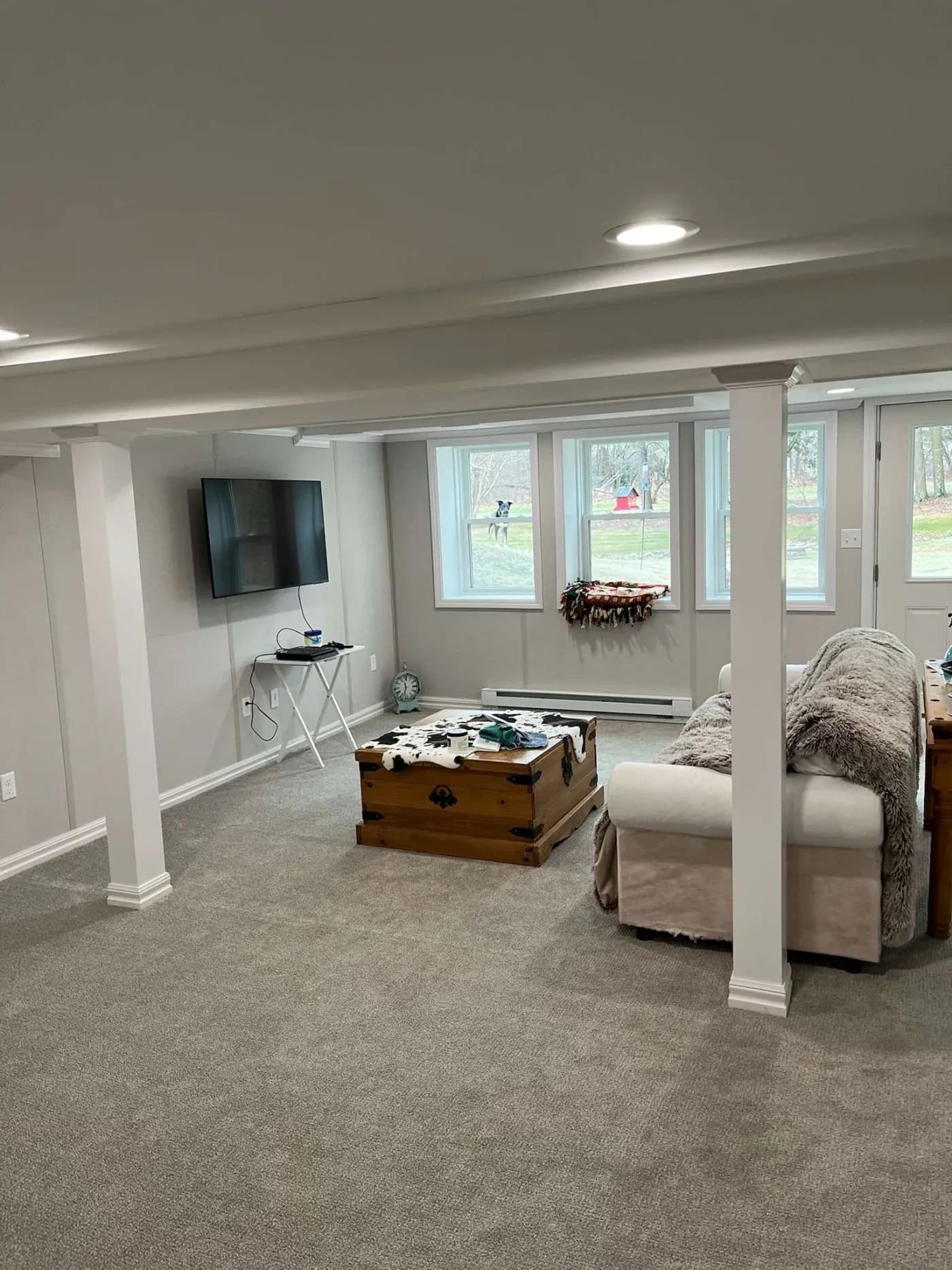Cozy basement living room with beige carpets, a soft cream couch with gray throw, chest coffee table, TV mounted on the wall, and large windows offering a garden view.