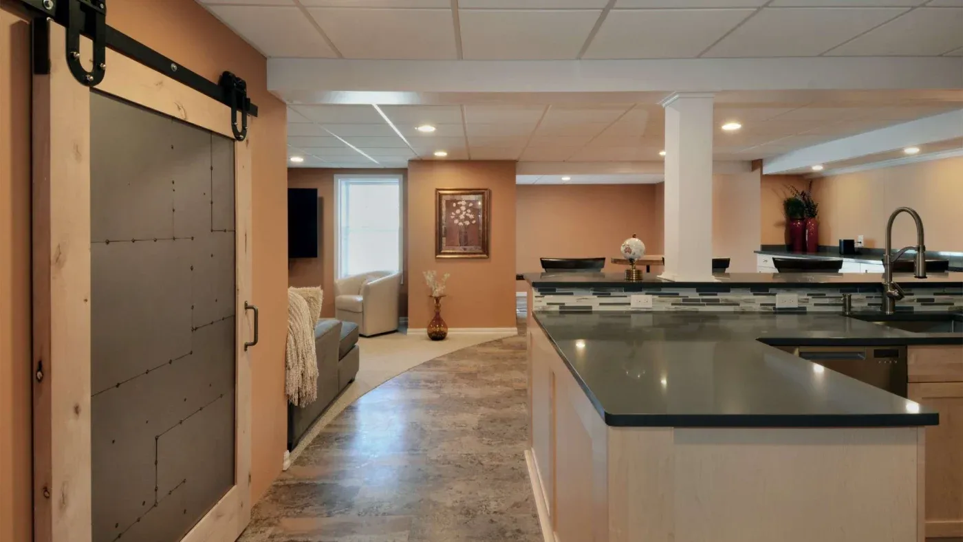 Modern basement with beige walls and polished floors, featuring a sliding barn door, cozy seating area, artwork, and a sleek kitchenette with a tiled backsplash.