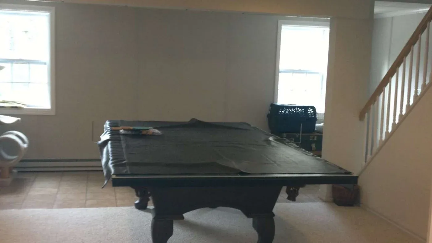A pool table covered with a dark cloth stands in a small, well-lit room with neutral walls. Two windows on either side enhance the airy ambiance.
