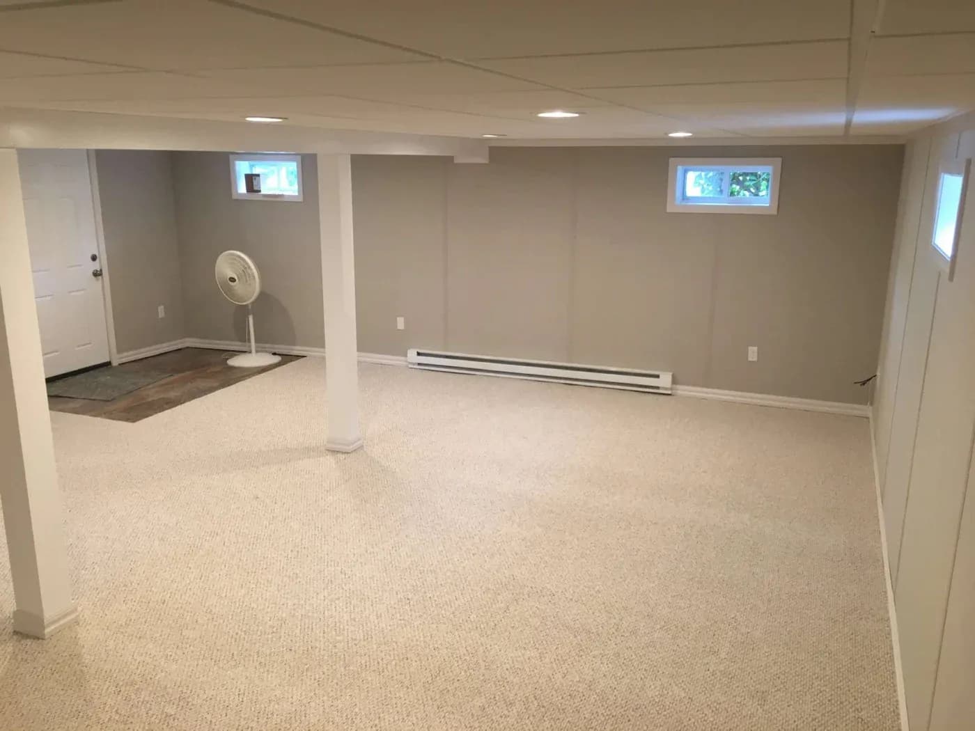 A spacious, beige-carpeted basement with white walls and ceiling, featuring small rectangular windows, a standing fan, and a closed door in the corner.