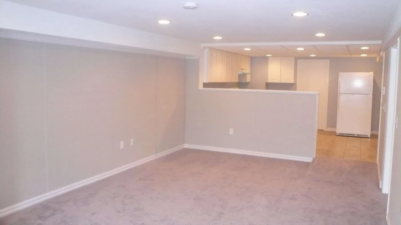 A spacious, empty basement features neutral walls, soft lighting, and carpet flooring. A small kitchen with white cabinetry and a fridge is visible.