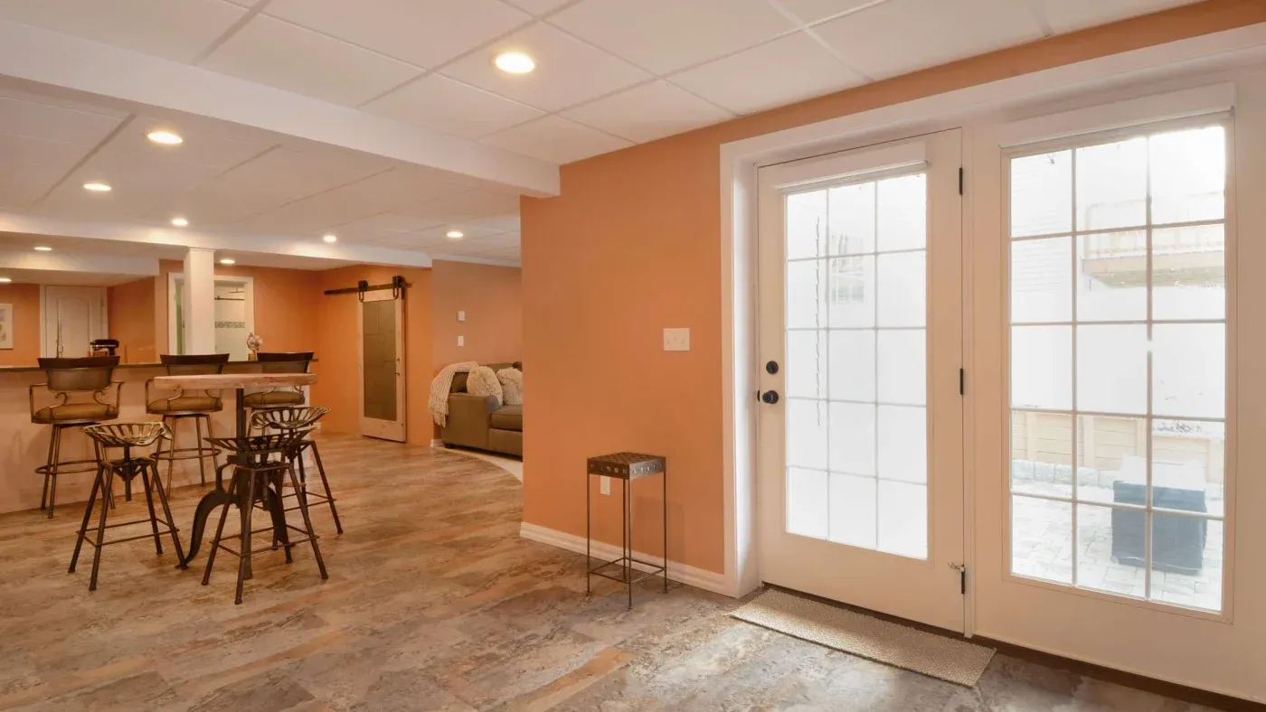 Spacious basement with peach walls and wood flooring, featuring a bar with stools, a cozy sofa, and double glass doors that let in ample natural light.