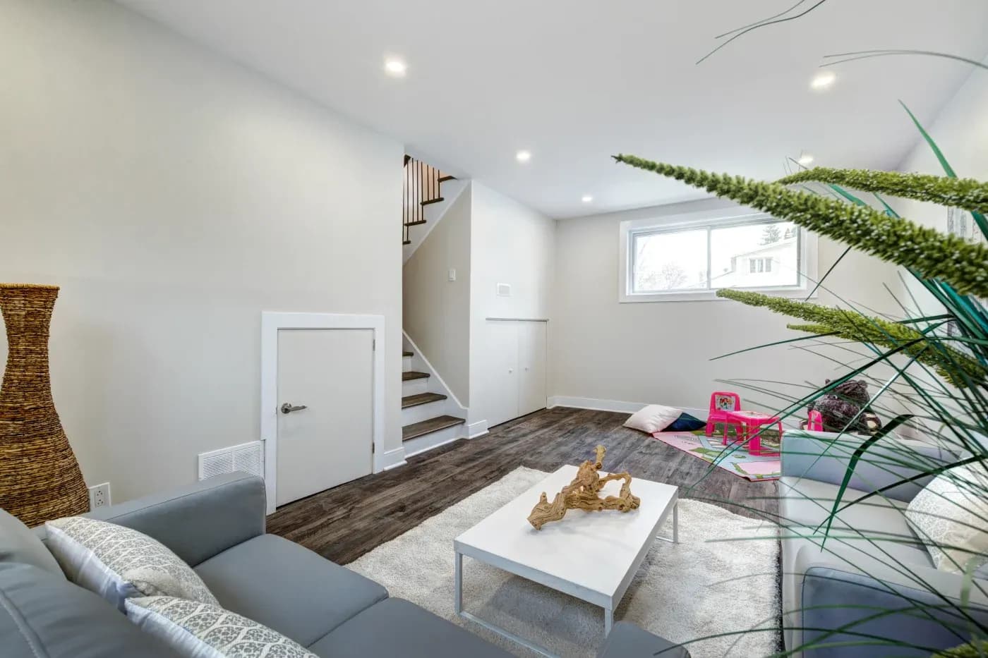 Bright modern living room with gray couches, a white coffee table, and wooden flooring. A staircase, plants, and children's toys add warmth and liveliness.