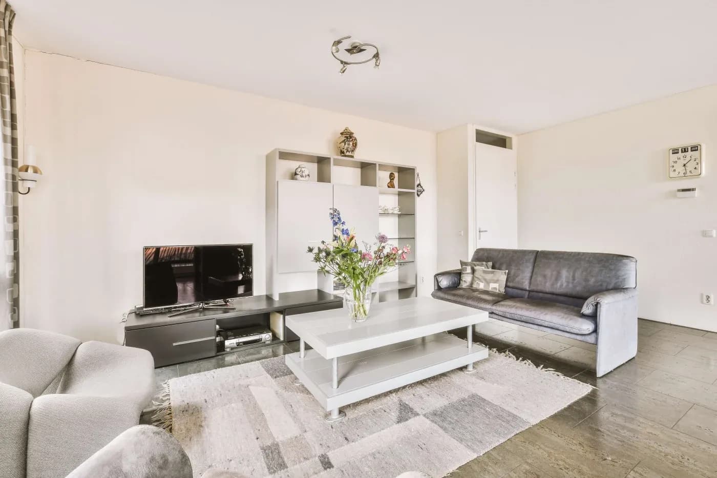 A modern living room with a gray couch and armchair, white coffee table with flowers, TV, and shelves with decor against a cream wall, creating a cozy ambiance.