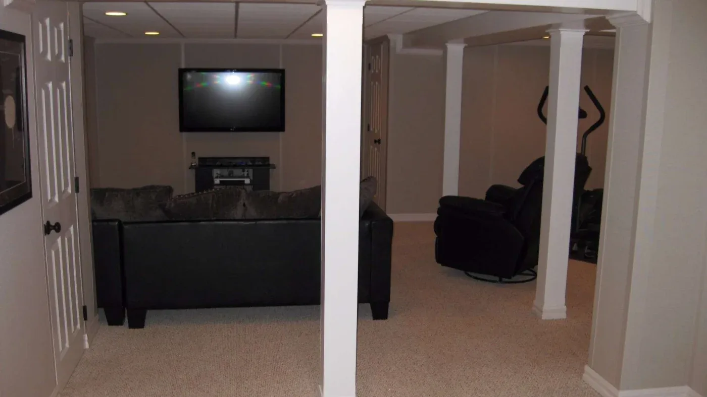 A cozy basement room with a TV on the wall, a black couch facing it, beige carpet, and a recliner with an exercise bike in the corner, lit softly.