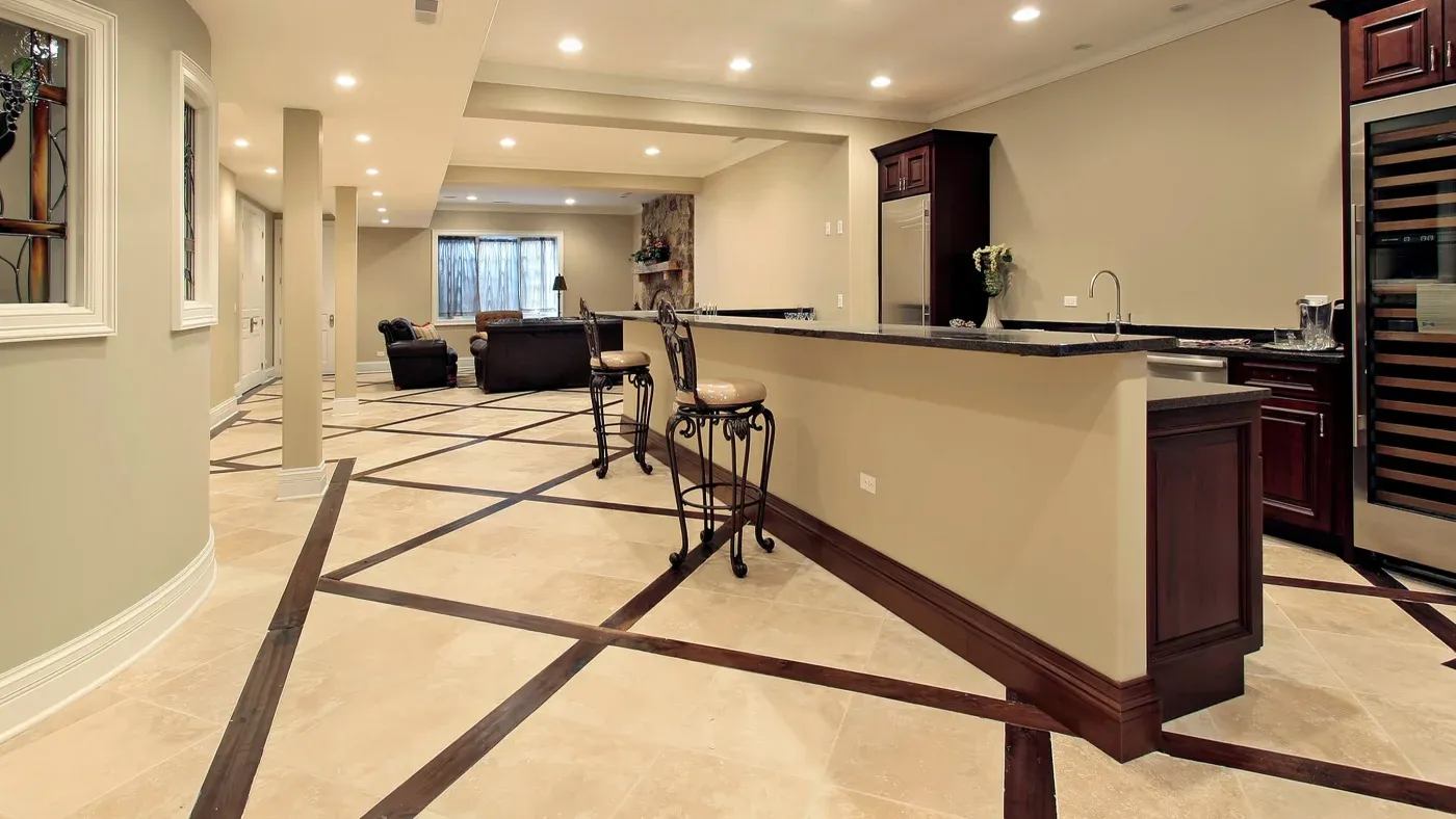 Spacious modern basement with beige walls and geometric patterned tiles. Features a long bar, elegant chairs, and a cozy seating area with a fireplace.