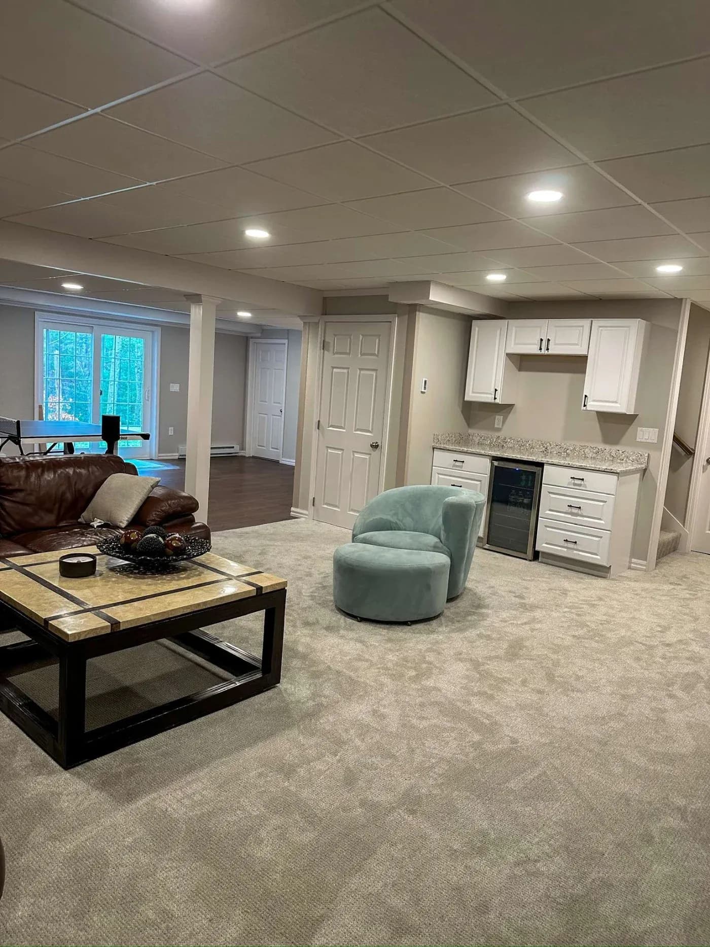 Spacious basement with beige carpet, white cabinets, and a small fridge. A cozy seating area features a plush blue chair and a brown leather sofa.