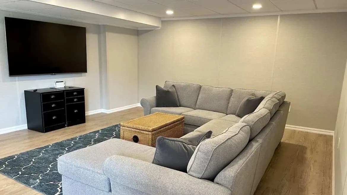 A cozy living room with a large gray sectional sofa, a wicker coffee table, and a patterned rug on wooden floors. A flat-screen TV is mounted on the wall.