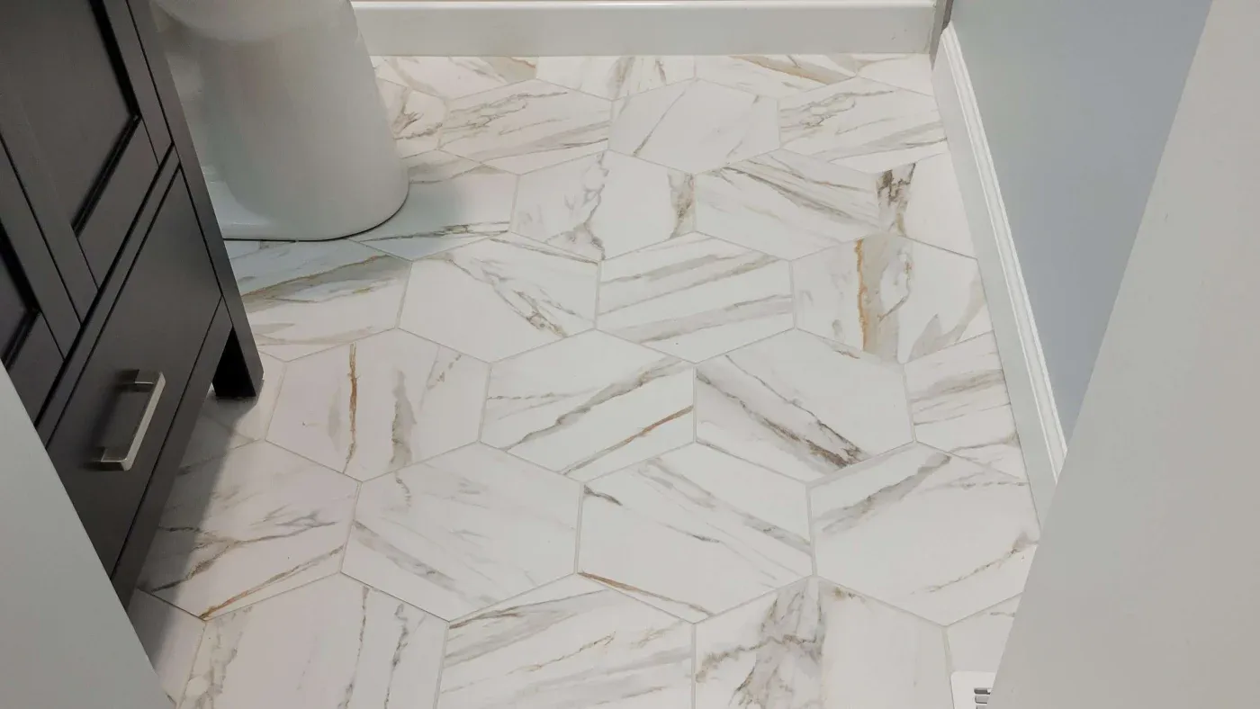 Hexagonal white marble tiles with subtle gray and brown veining on a bathroom floor, adjacent to a dark wooden vanity and white toilet.