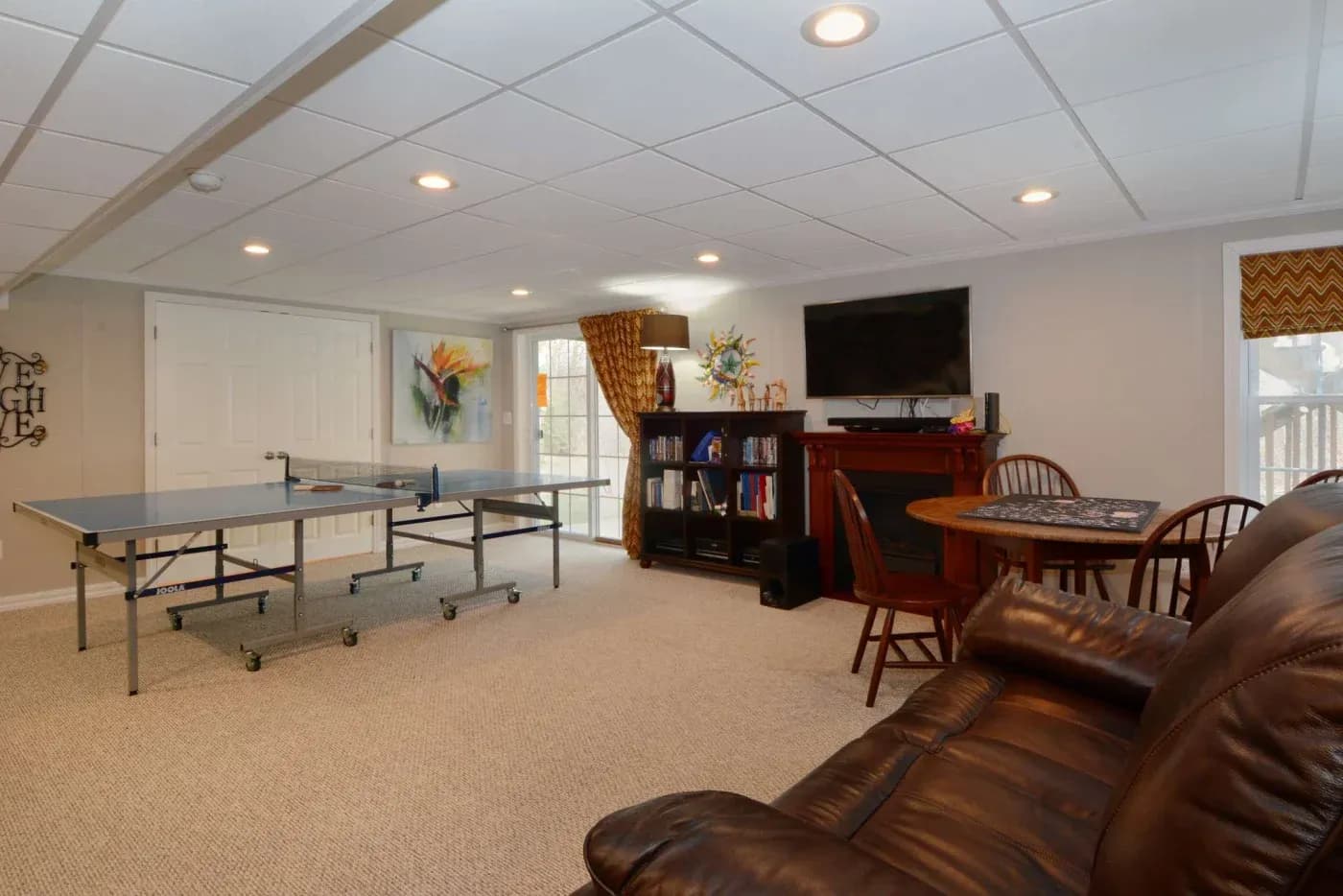 Spacious basement with brown leather couch, ping pong table, TV above a fireplace, bookshelves, and a round wooden dining table with chairs. Cozy ambiance.