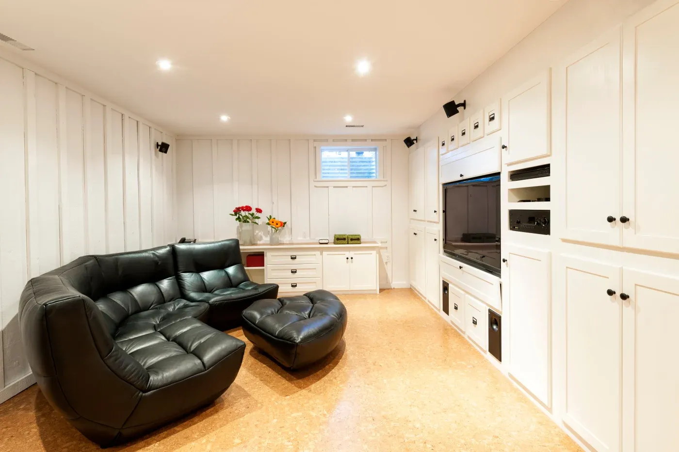 Cozy basement living room with a black leather sectional sofa and ottoman. White paneled walls, built-in cabinets, and a flat-screen TV create a modern, inviting atmosphere.