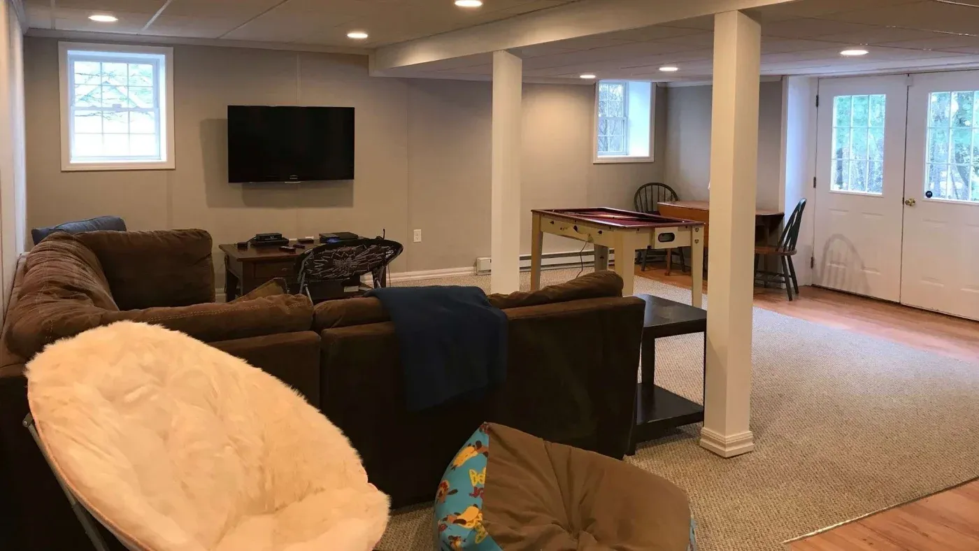 Spacious basement room with brown sectional sofa, wall-mounted TV, foosball table, and cozy chairs. Bright natural light from large windows.