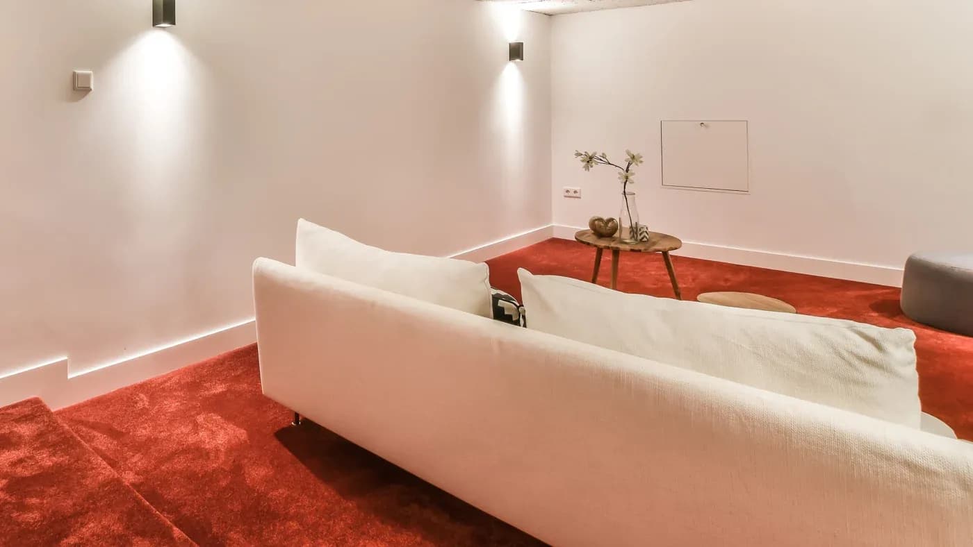 Modern living room with a white sofa on red carpet, illuminated by wall lights. A small wooden table with decor adds a cozy touch.