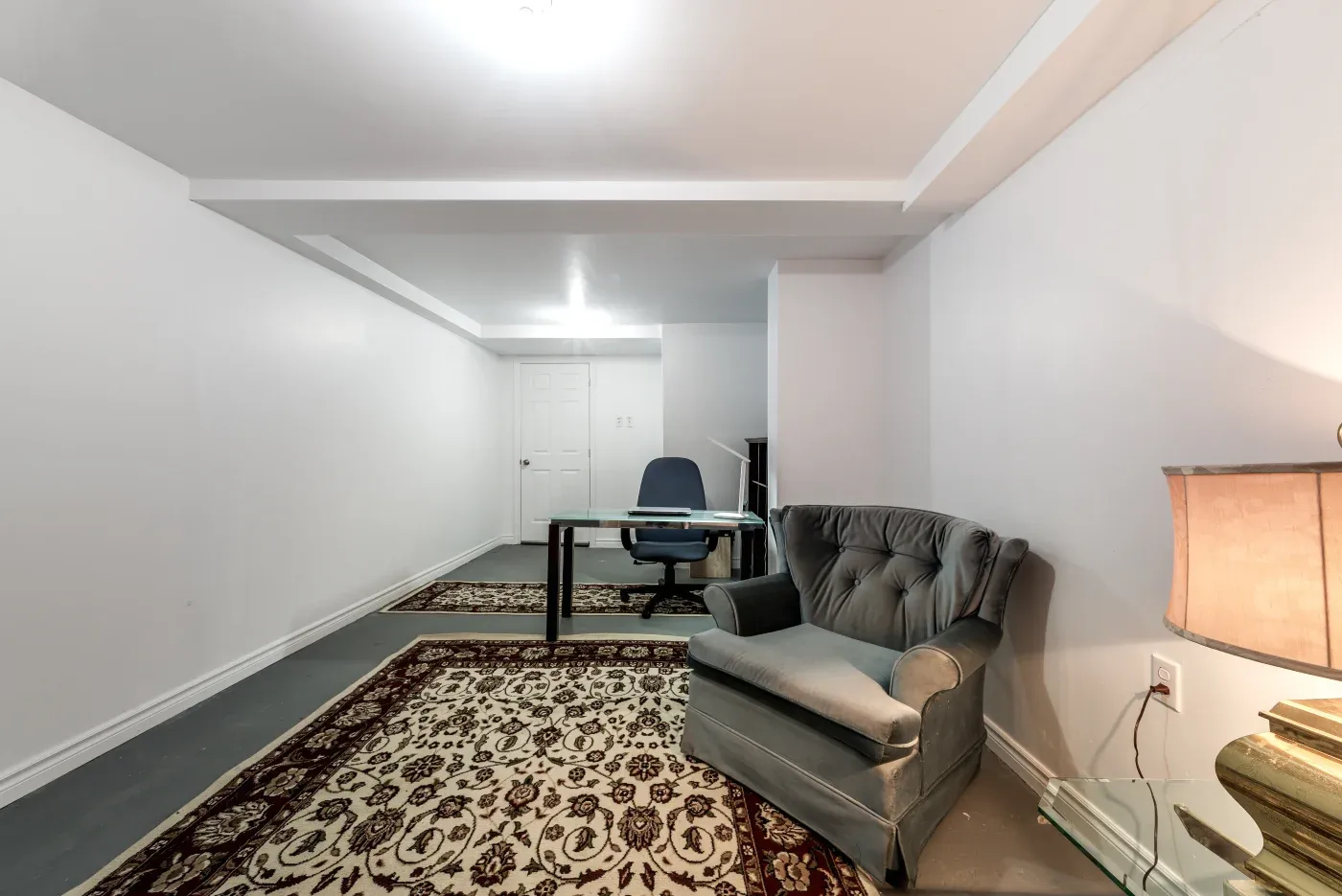 Spacious basement room with white walls, patterned rugs, and a glass desk with a blue chair. A cozy grey armchair and a lit table lamp add warmth to the space.