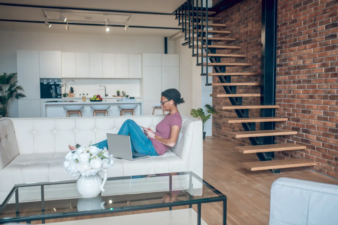 Woman in a relaxed modern loft with brick walls and wooden stairs, seated on a white sofa, using a laptop, conveying a calm, productive atmosphere.