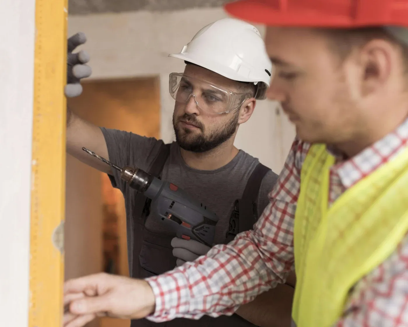 Two construction workers, wearing hard hats and safety gear, collaborate on a building project. One uses a drill, focused and engaged.