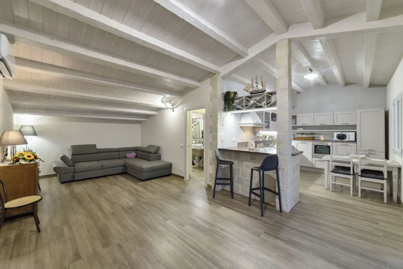 Spacious open-plan living area with wooden floors and white-beamed ceiling. Features a large gray sectional sofa, modern kitchen, and a cozy dining area.