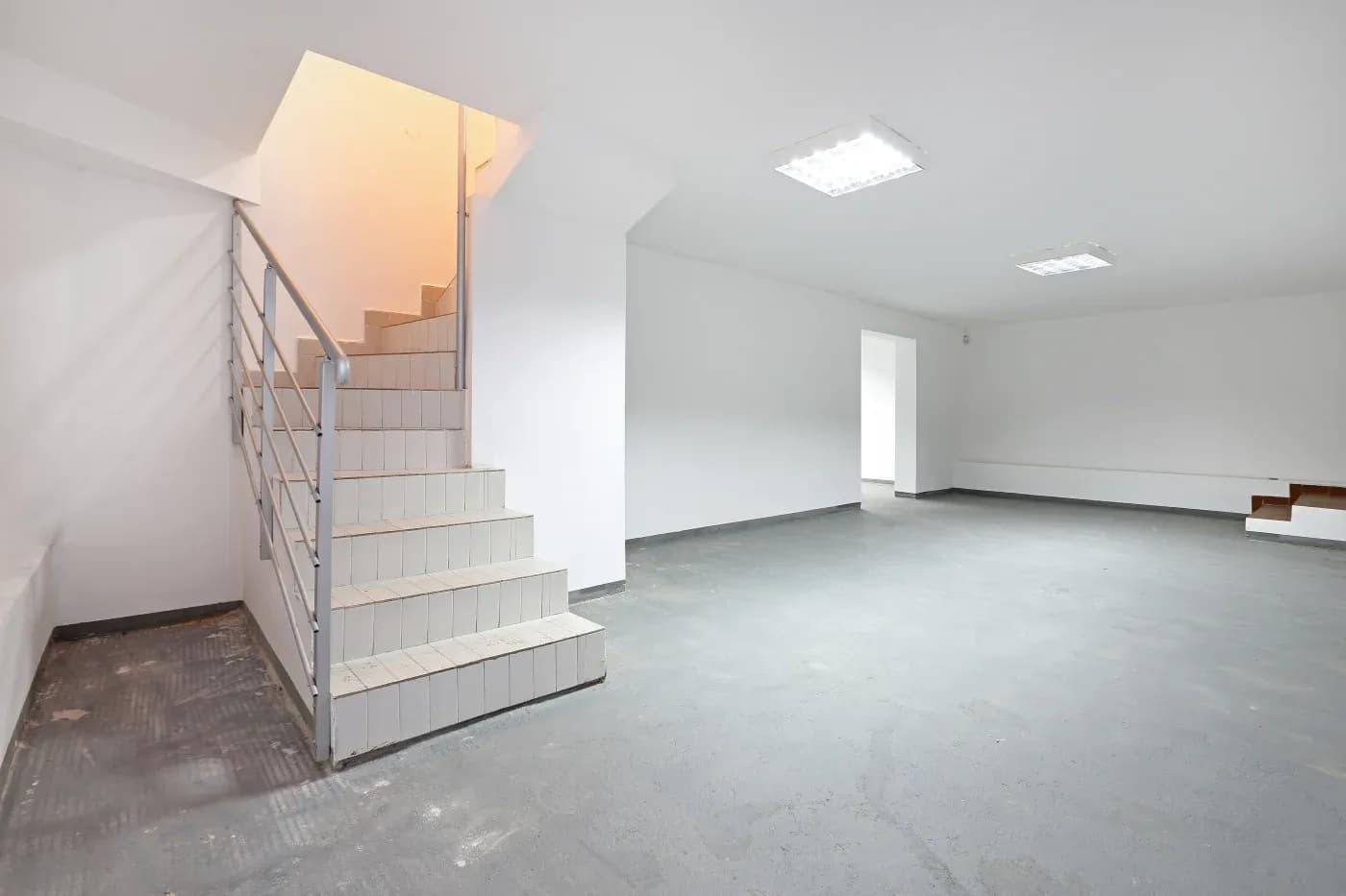 Spacious, empty basement with white walls, gray floor, and fluorescent lights. A staircase with metal railing leads to an upper floor. Minimalist and clean.
