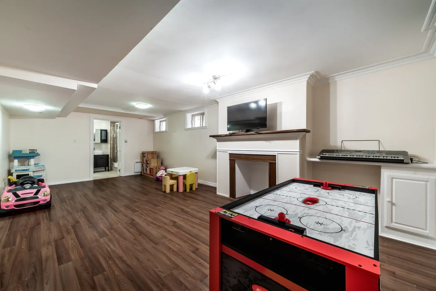 Spacious basement playroom with wood flooring, featuring an air hockey table, toy car, small children’s table, TV, and keyboard, creating a playful atmosphere.