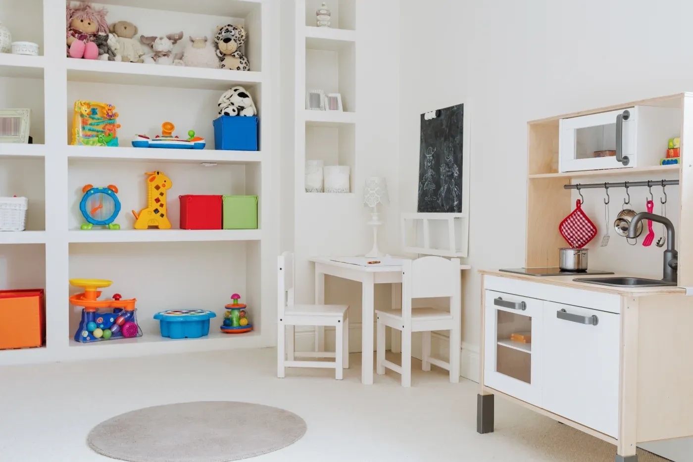 Bright playroom with white shelves filled with colorful toys and stuffed animals. A small wooden kitchen set and a child-sized table create a cozy space.