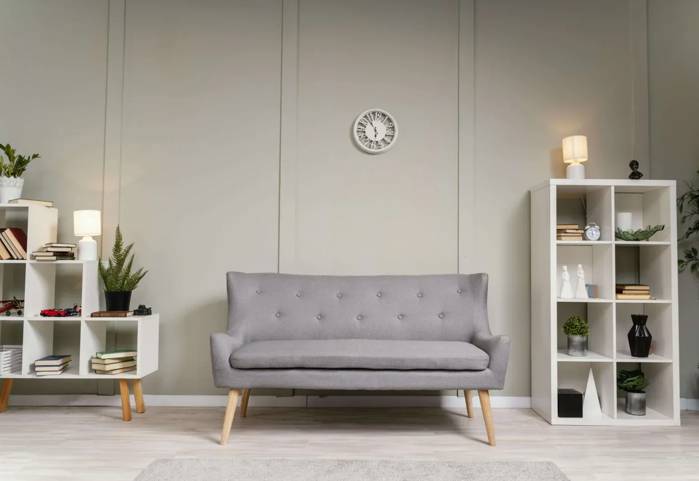 Modern living room with a gray sofa centered, flanked by white bookshelves filled with books and plants. A clock hangs on the wall above the sofa. Cozy lighting.