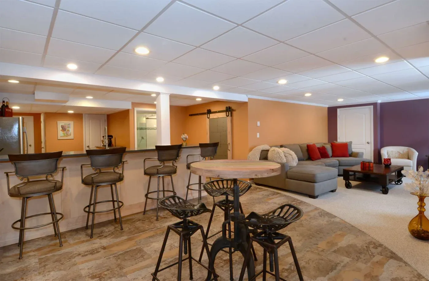 Spacious basement with a modern bar setup, high stools, rustic round table, and cozy sectional sofa adorned with red pillows. Warm lighting creates an inviting vibe.