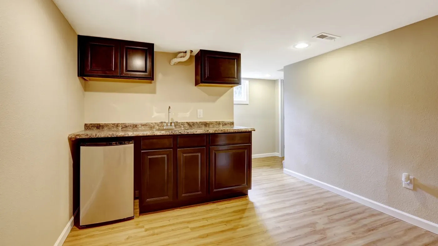 A small, modern kitchenette with dark wood cabinets, a granite countertop, and a stainless steel mini-fridge against beige walls and light wood flooring.