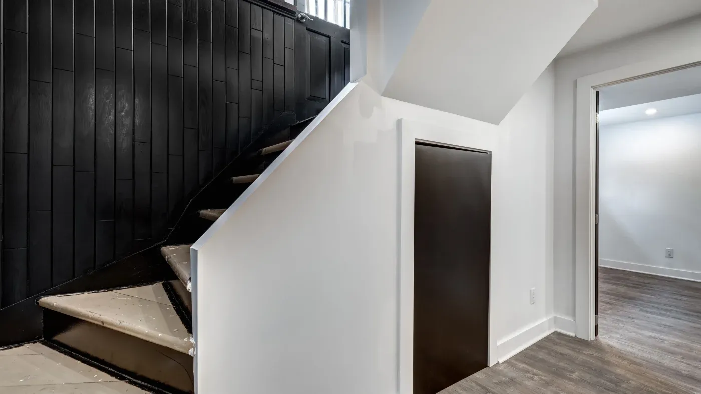 A modern interior space featuring a black wooden staircase against a white wall. The room has dark wood flooring and an open doorway, creating a sleek and minimalist atmosphere.