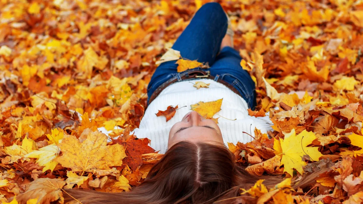 A person in a white sweater and jeans lies on colorful autumn leaves, evoking a sense of relaxation and the beauty of fall.