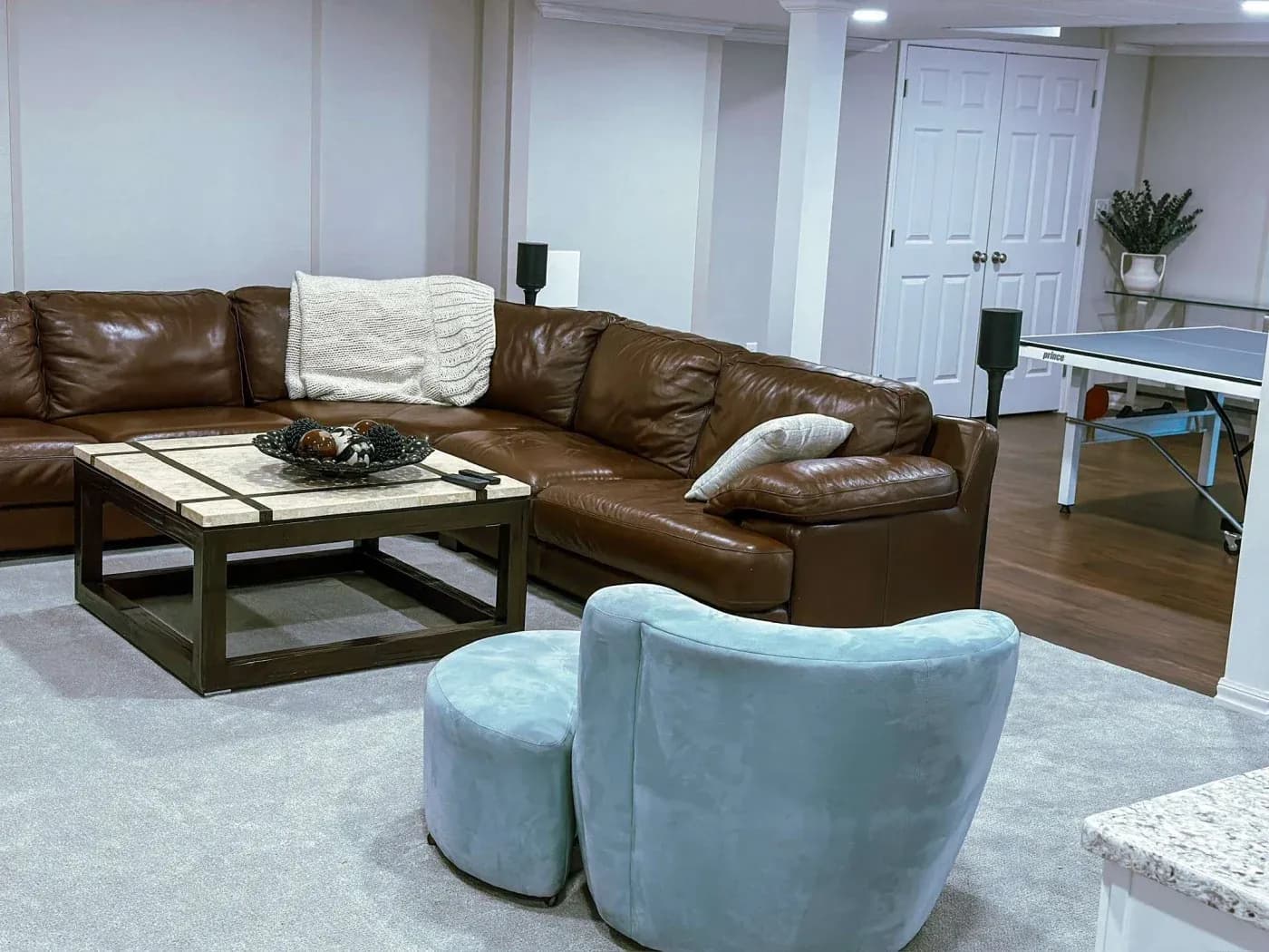 A cozy basement living area features a brown leather sectional, a beige throw, a pale blue chair, and a tiled coffee table. A ping pong table is visible.