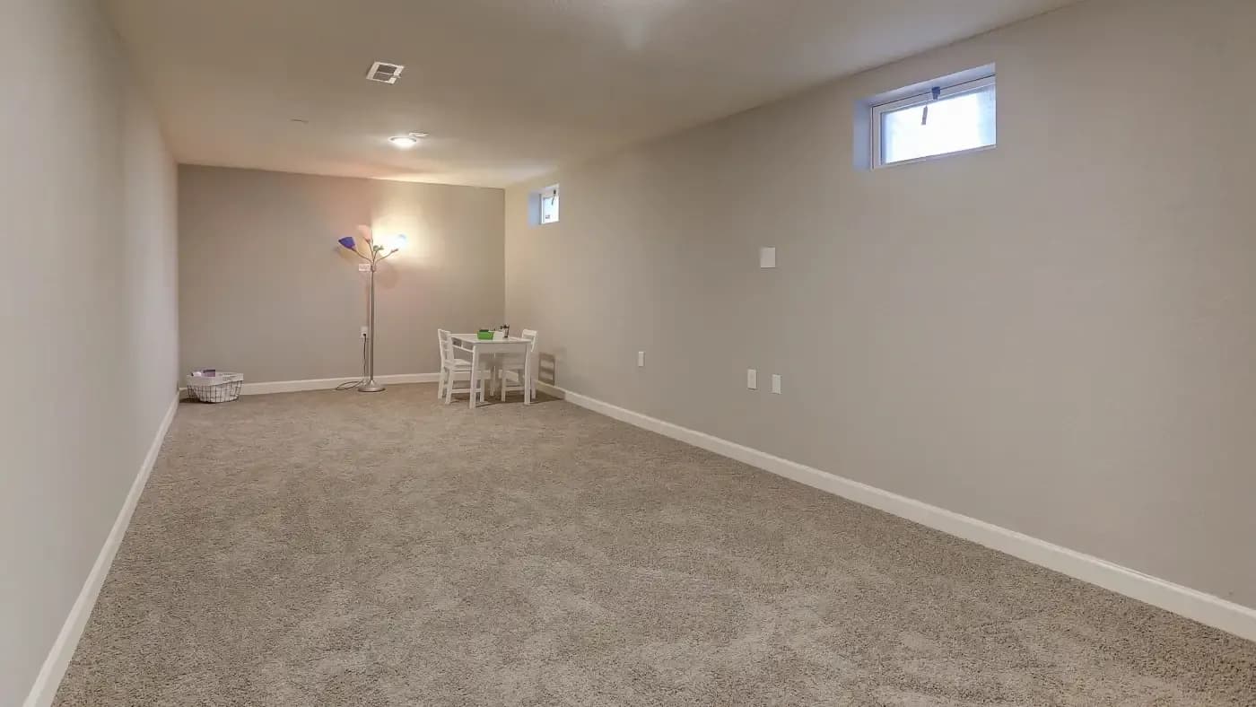 A spacious, carpeted basement room with neutral walls, featuring a small table and chairs, a colorful floor lamp, and small windows. The ambiance is calm and minimalistic.