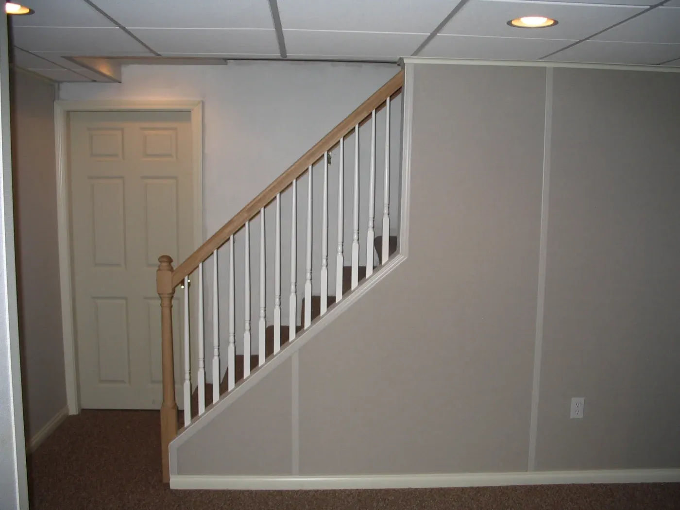 A basement with beige walls features a wooden staircase with white railings. A closed door is on the left, and the space is lit by recessed lights.
