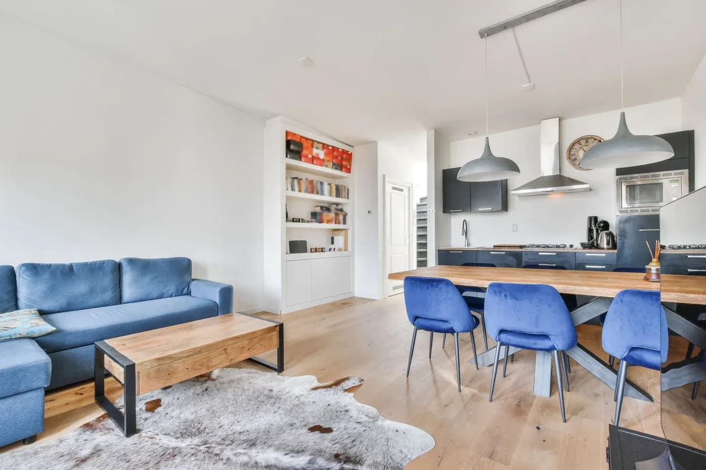 A modern living and dining area with light wood floors, a blue sofa, cowhide rug, wooden coffee table, blue dining chairs, and a sleek kitchen.