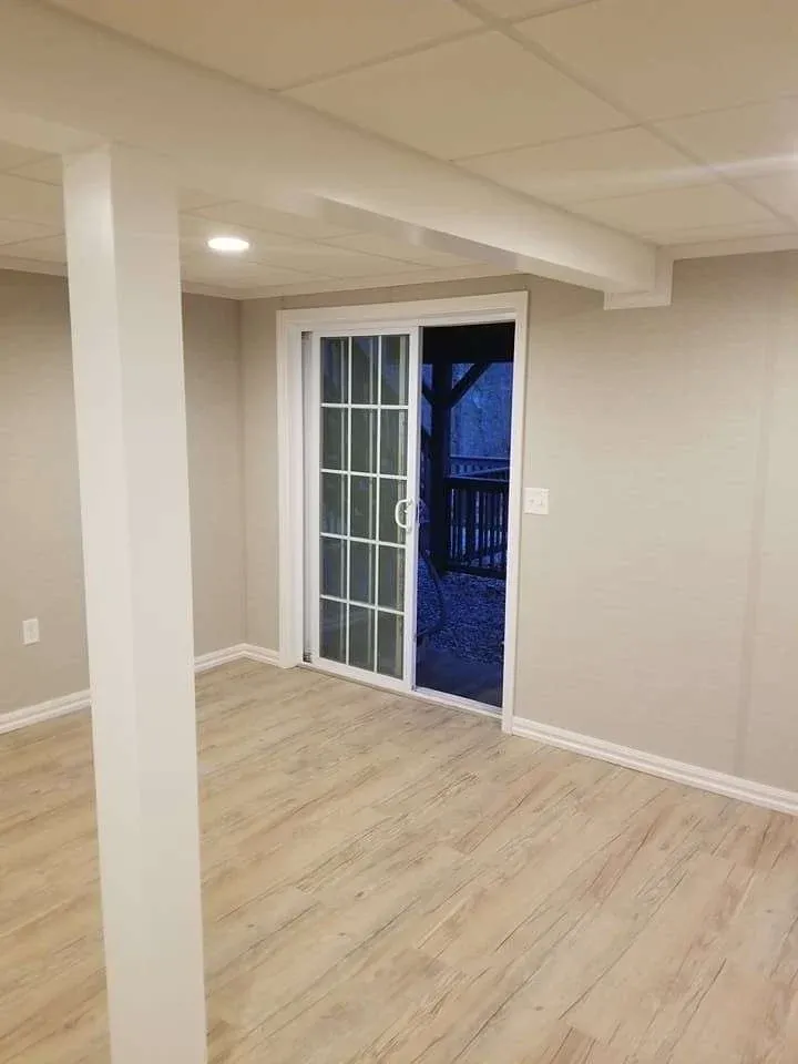 A bright, empty room with light wood flooring and beige walls. A sliding glass door opens to a dimly lit outdoor area, creating a peaceful atmosphere.