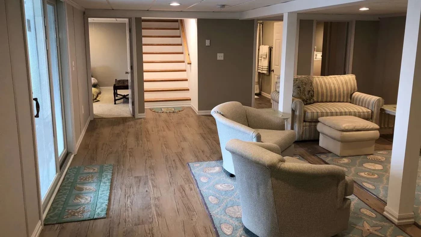 A cozy basement living area with wooden floors, sliding glass doors on the left, soft armchairs, a striped couch, and a staircase leading up in the center.