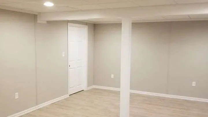 A minimalist, empty basement room with beige walls, laminate flooring, a single support column, recessed lighting, and a white door, evoking a clean, spacious feel.