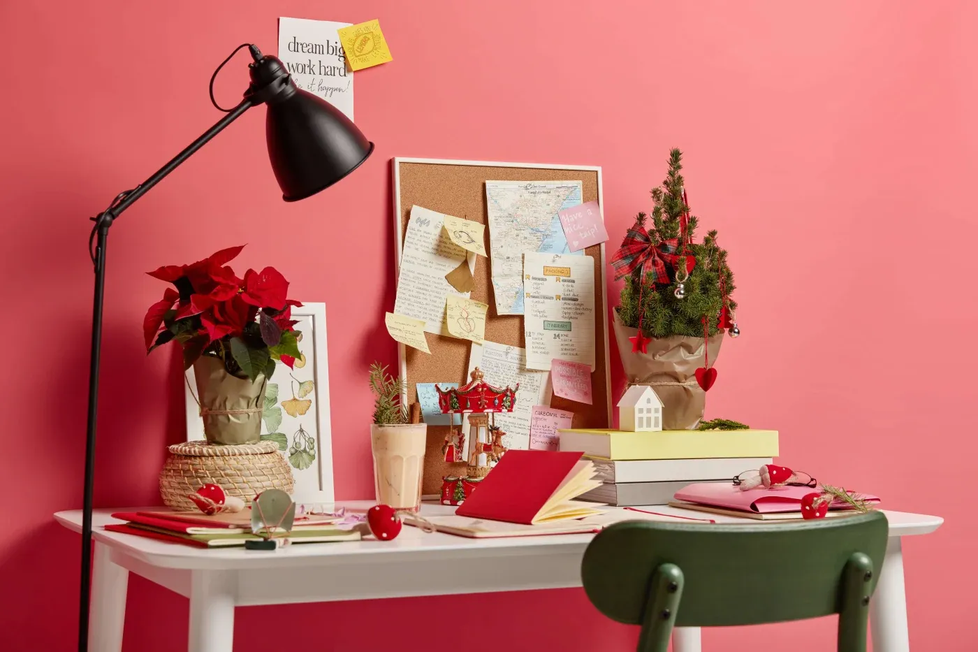 A cozy desk against a pink wall features a lamp, plants, a corkboard with notes, and a festive mini tree, evoking a creative, cheerful atmosphere.