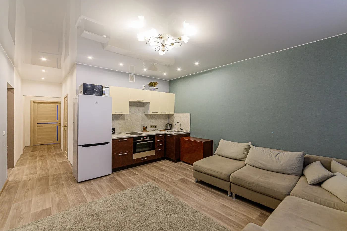Modern living room with open kitchen. Beige sofa, light wood flooring, and teal accent wall create a cozy atmosphere. Overhead chandelier adds warmth.