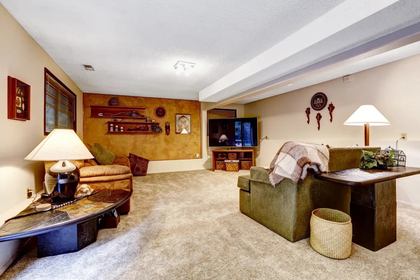 Cozy basement with beige carpet, warm lighting, and earth-toned walls. Features a plush sofa, rustic decor, shelf, and TV, creating a relaxing ambiance.