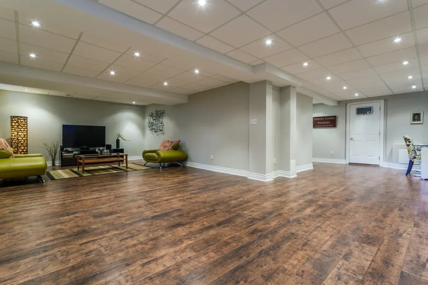 Spacious basement with warm wooden floors, neutral walls, and recessed lighting. Features a TV area with green sofas, a floor lamp, and decorative art.