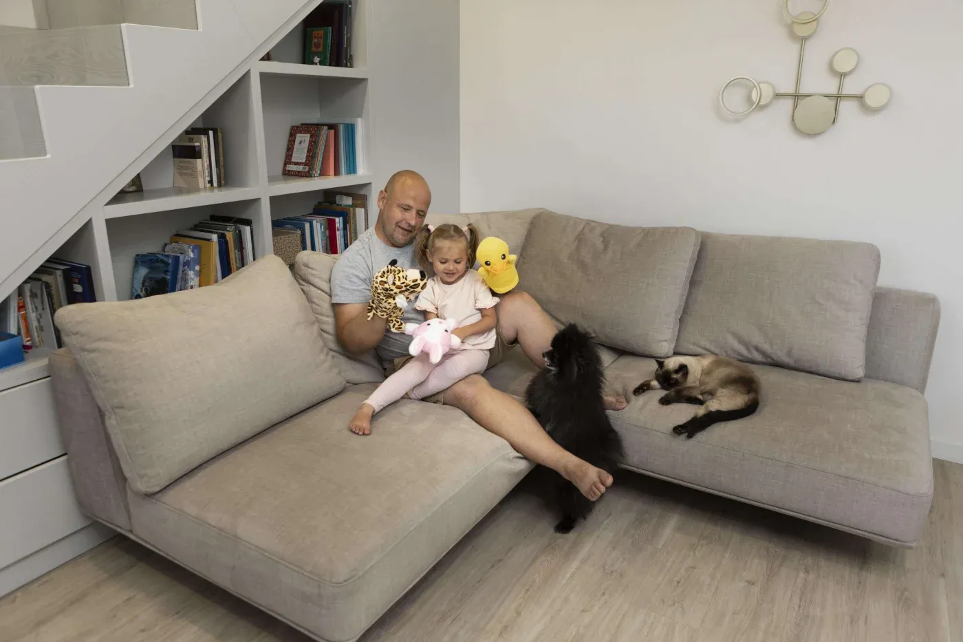 A man and child sit on a gray sofa in a cozy living room, holding plush toys. A black dog and a Siamese cat relax nearby. The scene feels warm and cheerful.