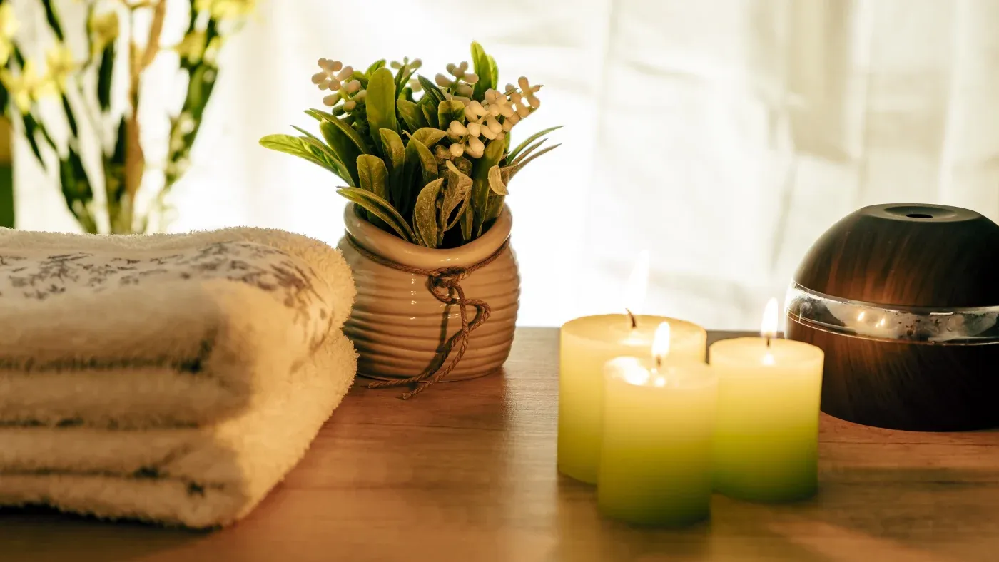 A serene spa setting with glowing candles, a small green plant in a pot, and folded towels on a wooden surface, conveying relaxation and warmth.