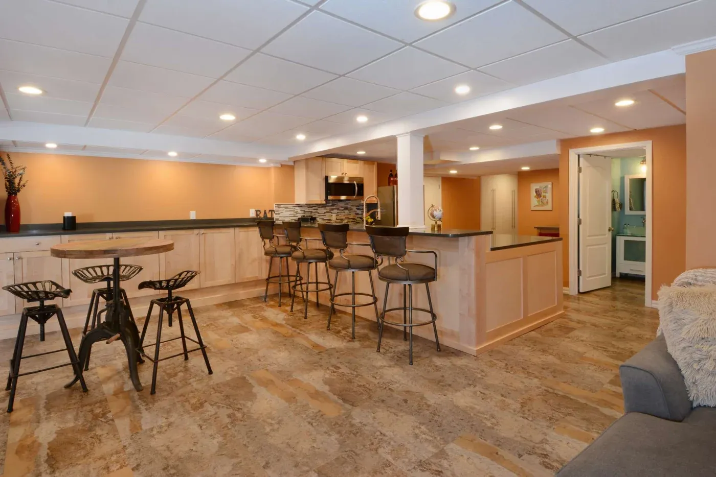 Spacious basement bar area with warm lighting, wood floors, and peach walls. Features a long counter with five barstools and a small table with four chairs.