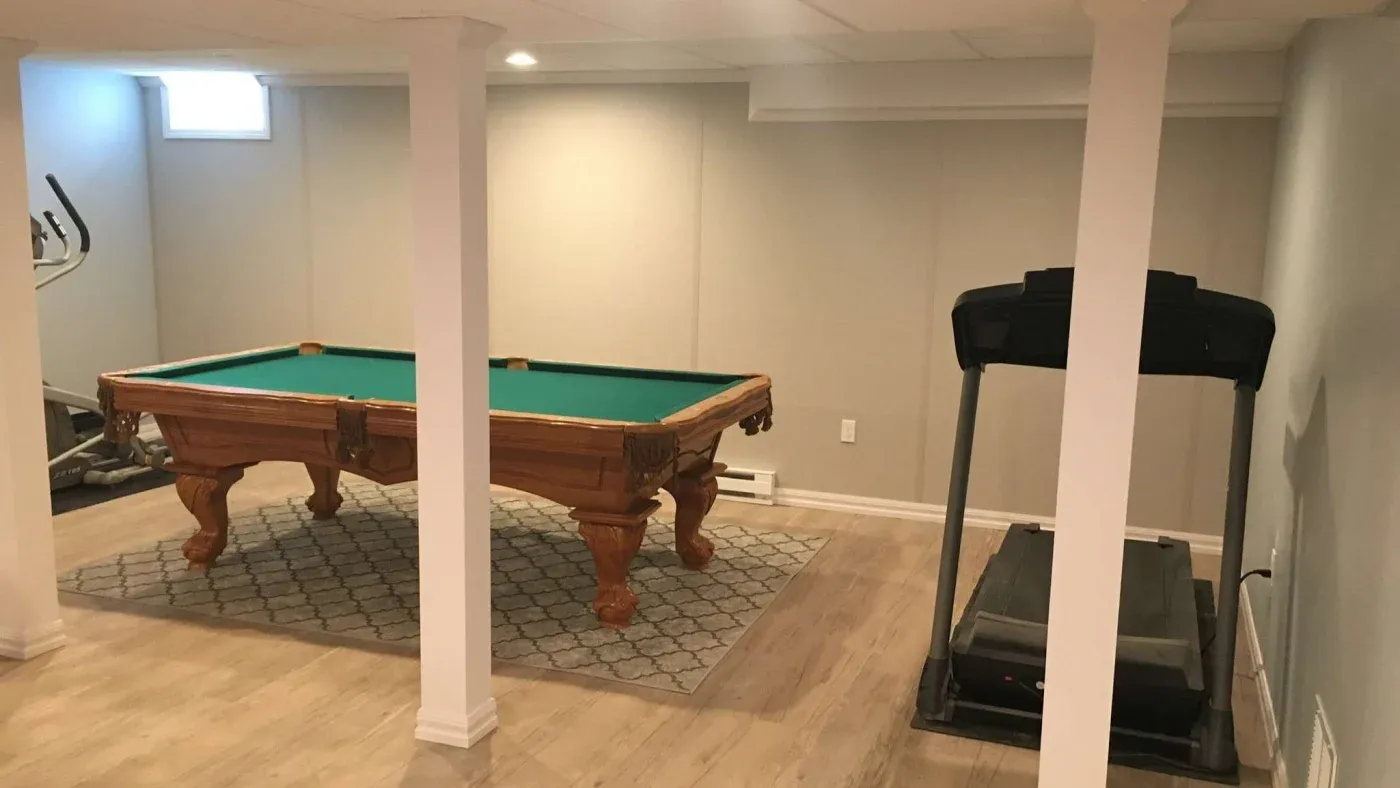 A cozy basement with a billiard table on a patterned rug and a treadmill on a wooden floor. Soft lighting and neutral walls create a calm atmosphere.