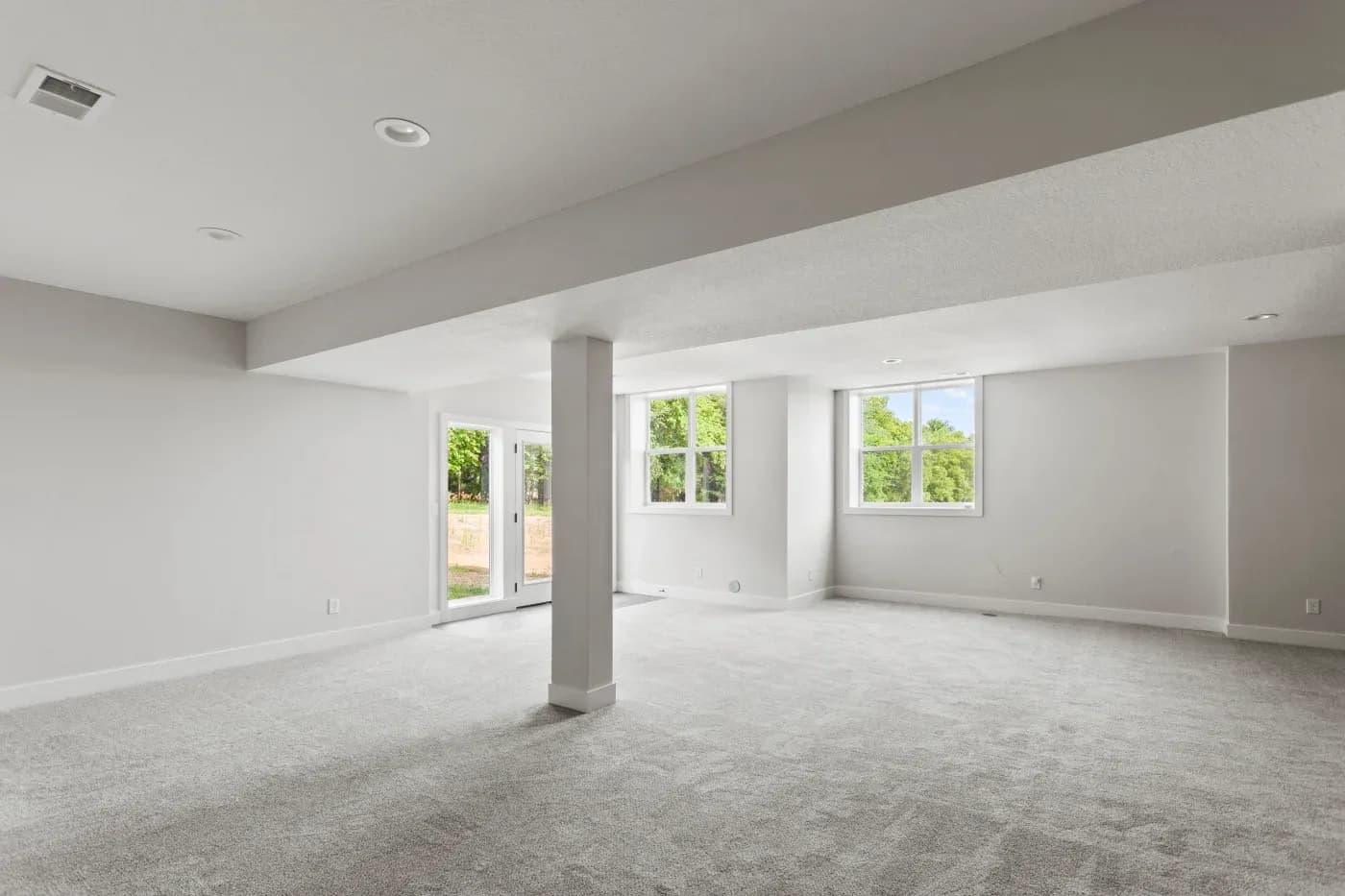 A spacious, empty room with light gray carpet and white walls, features large windows and a glass door. Sunlight streams in, creating a bright, open atmosphere.
