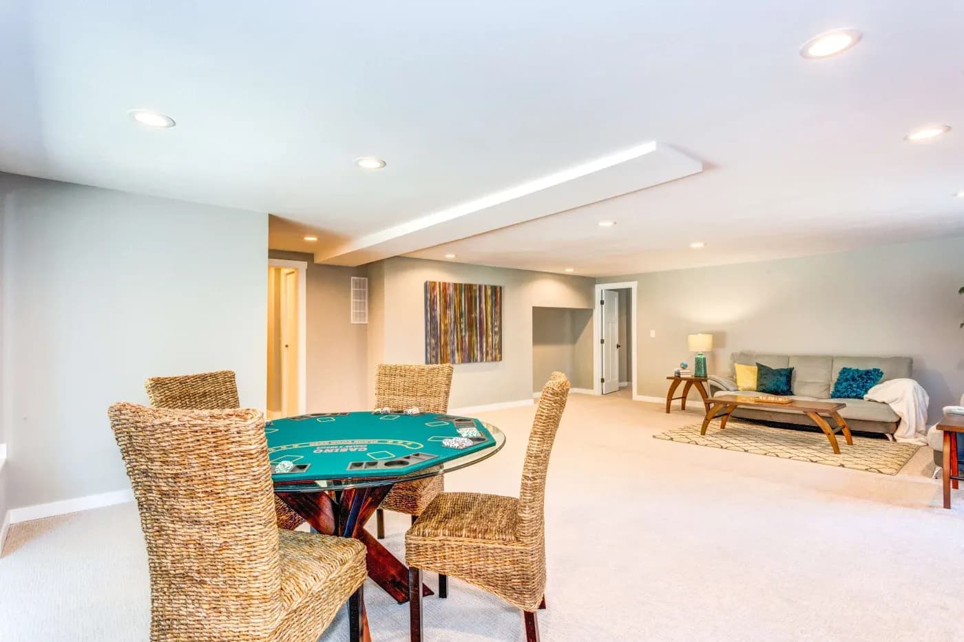 Spacious basement with a poker table, wicker chairs, beige carpet, and a cozy seating area with a sofa. Warm lighting and abstract art create a relaxed atmosphere.