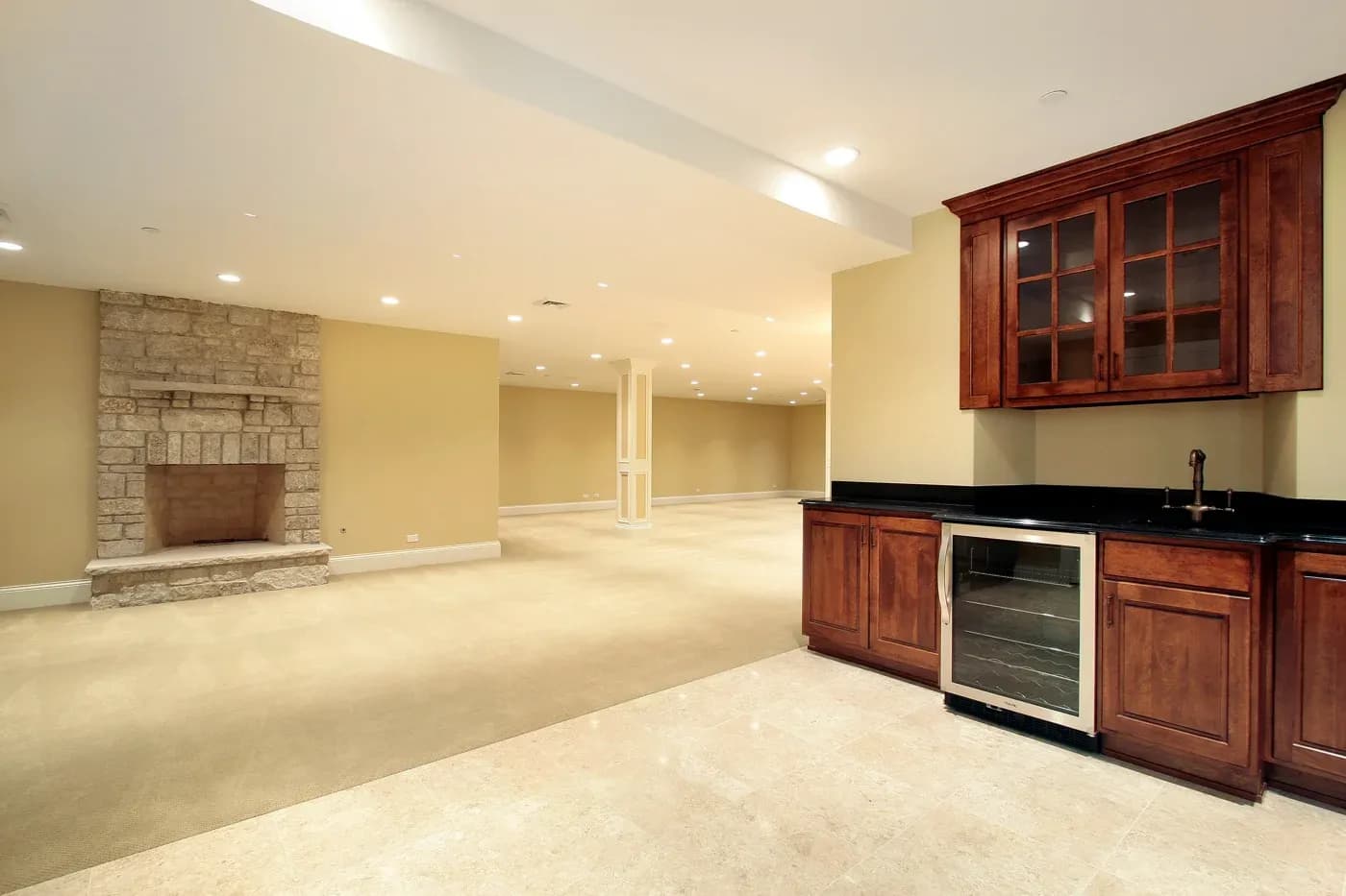 Spacious basement with beige walls, stone fireplace, and recessed lighting. Includes a wet bar with dark countertops and wooden cabinets. Cozy ambiance.