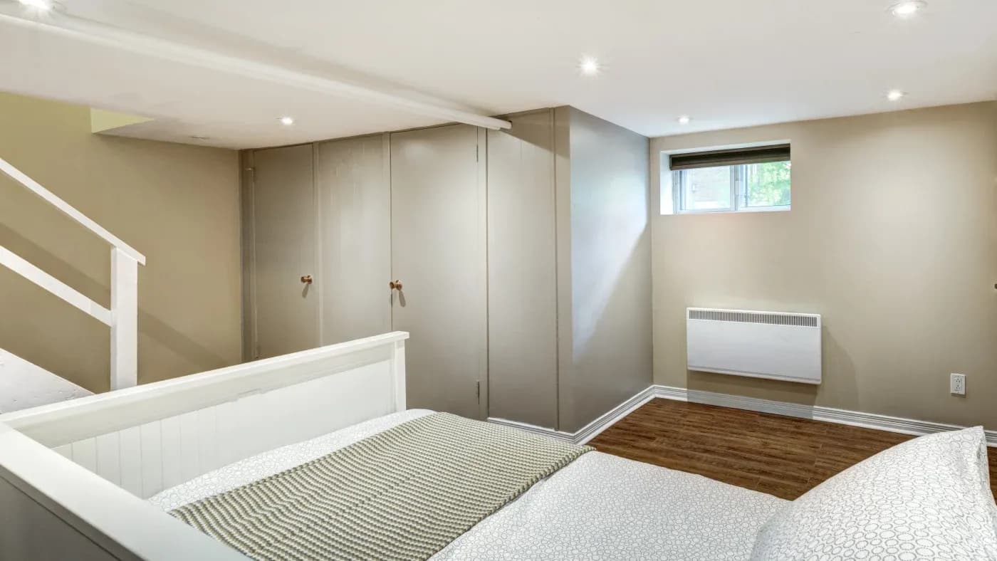 A minimalist bedroom with beige walls, white bedframe, patterned bedding, and wood floor. Soft lighting, small window, stairs, and a heater create a cozy feel.